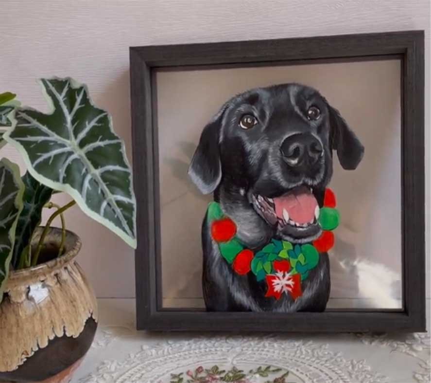 A custom pet portrait painted on clear acrylic, leaning against a wall in a sunlit corner with green indoor plants, showing the natural light reflecting on the painted surface.