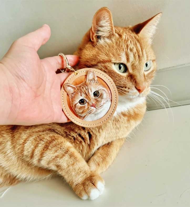 A hand holding a small custom pet memorial gift, a leather engraved tag with a cat portrait, positioned next to the real cat to show the detailed craftsmanship.