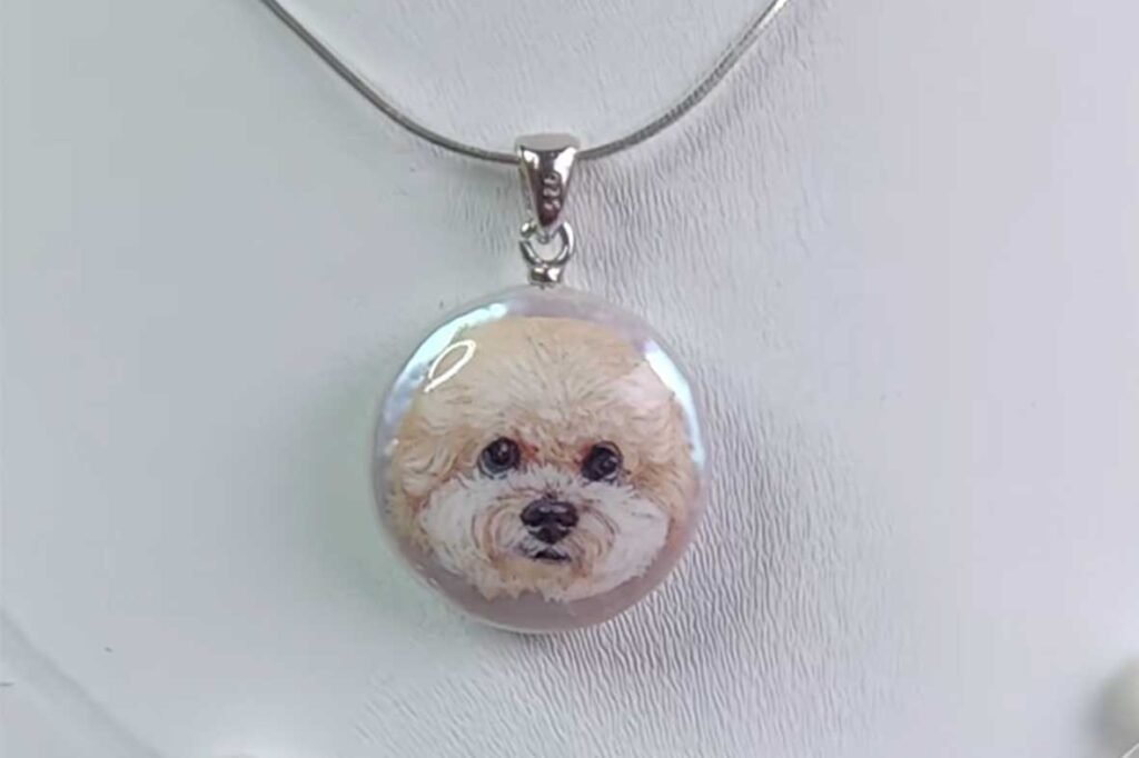 High-resolution close-up of a hand-painted brown puppy portrait on a pearl pendant against a white background.