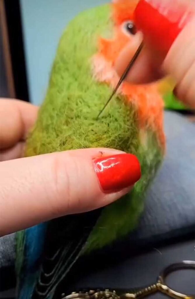 Artisan using a fine gauge needle to refine the green wool feathers and facial features of a needle-felted parrot.