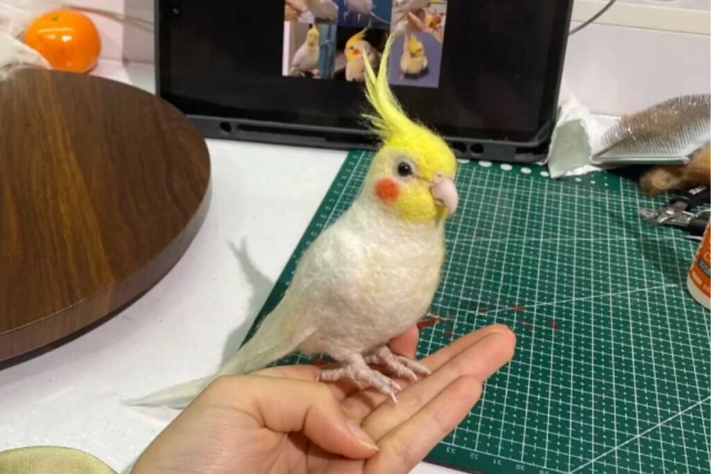 A finished white and yellow Cockatiel needle-felted sculpture standing on a human hand, with a workstation in the background.