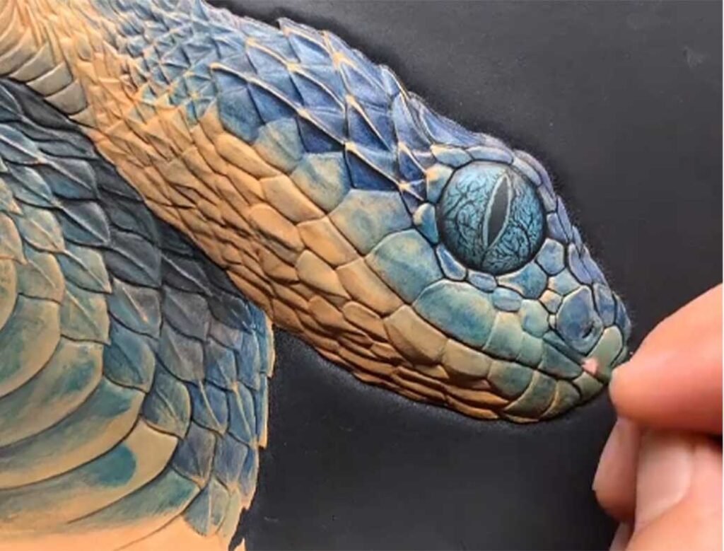Artisan hand-painting a hyper-realistic blue and yellow snake portrait on leather using a fine 0/3 liner brush to capture intricate scale details.
