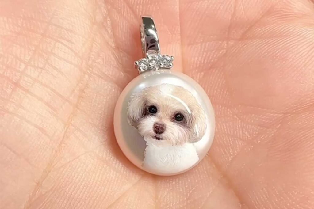 Close-up of a hand-painted white dog portrait on a pearl pendant held in a hand, showing intricate fur details.