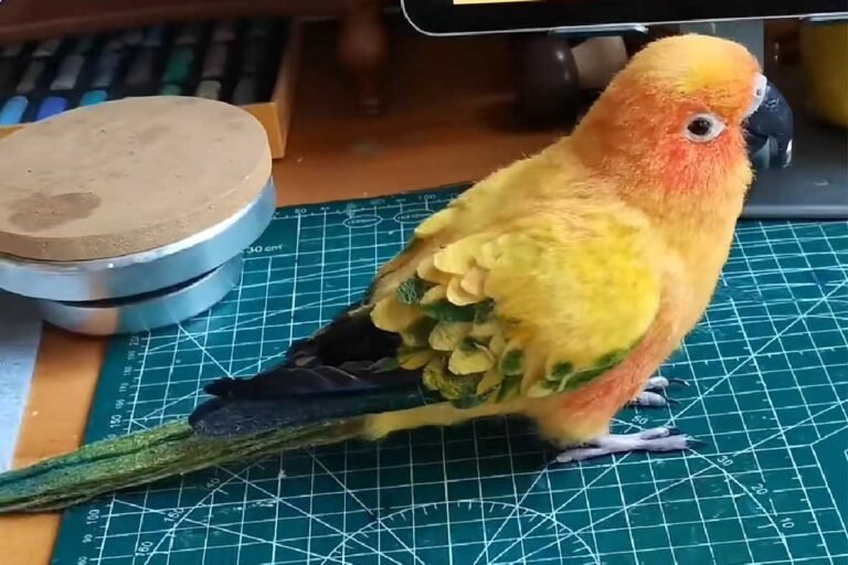 A hyper-realistic needle-felted Sun Conure parrot sculpture on a green cutting mat, showcasing intricate yellow and orange feather textures.