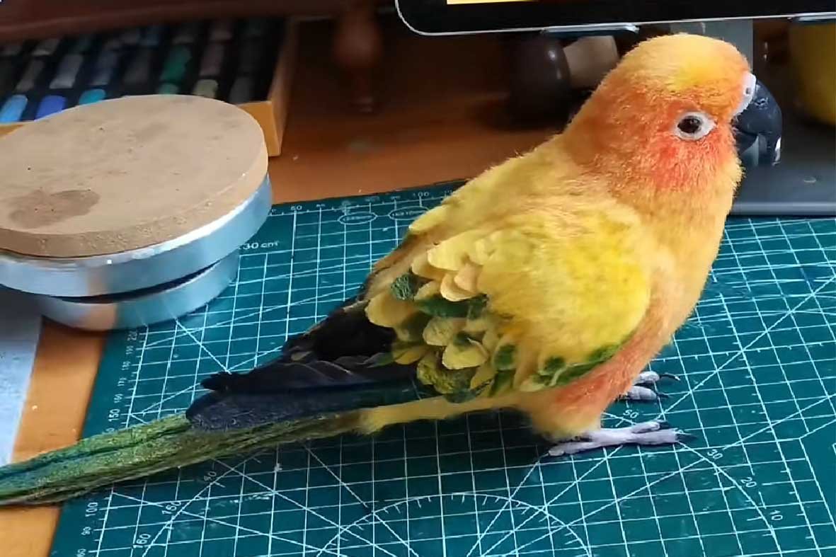 A hyper-realistic needle-felted Sun Conure parrot sculpture on a green cutting mat, showcasing intricate yellow and orange feather textures.
