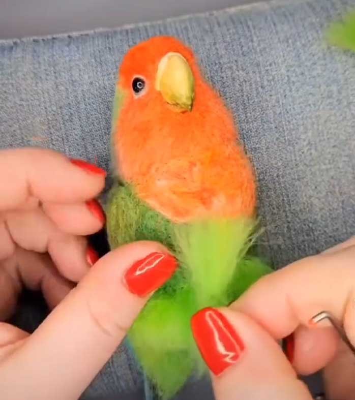 Close-up of artisan hands using a felting needle to shape the orange and green wool head of a parrot on a denim surface.