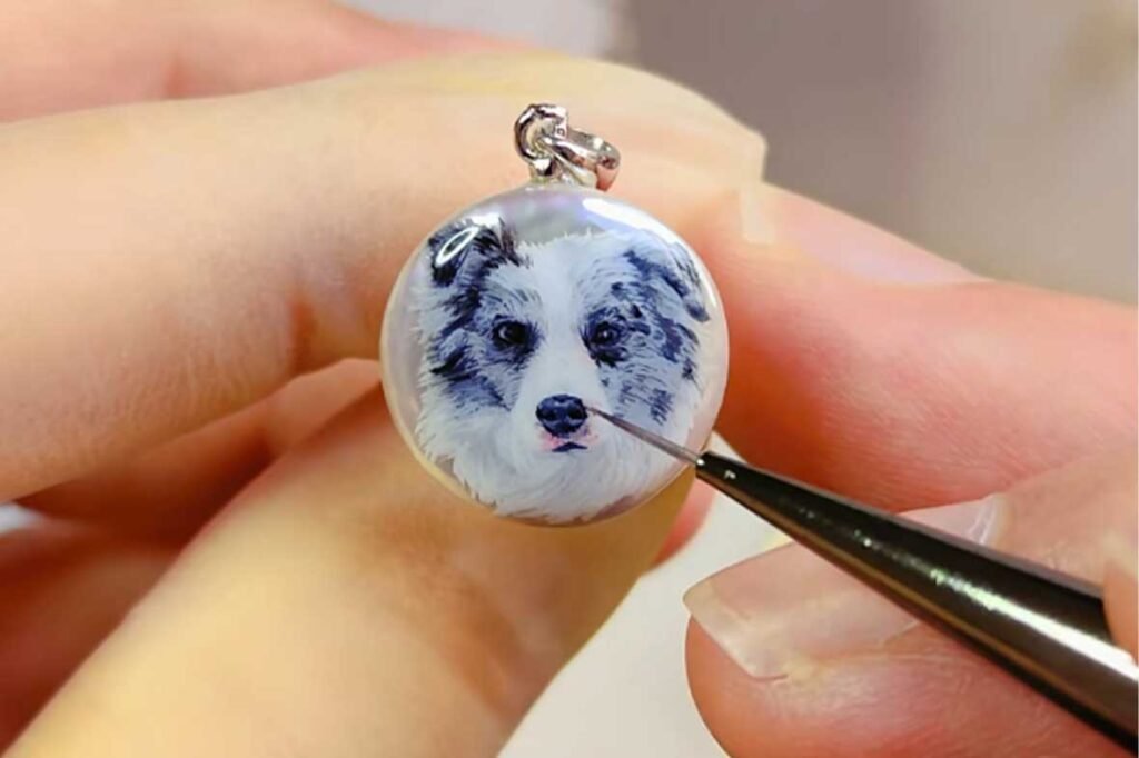 Macro shot of an artist using a fine-tipped brush to paint a Border Collie portrait on a 1.5cm pearl pendant.