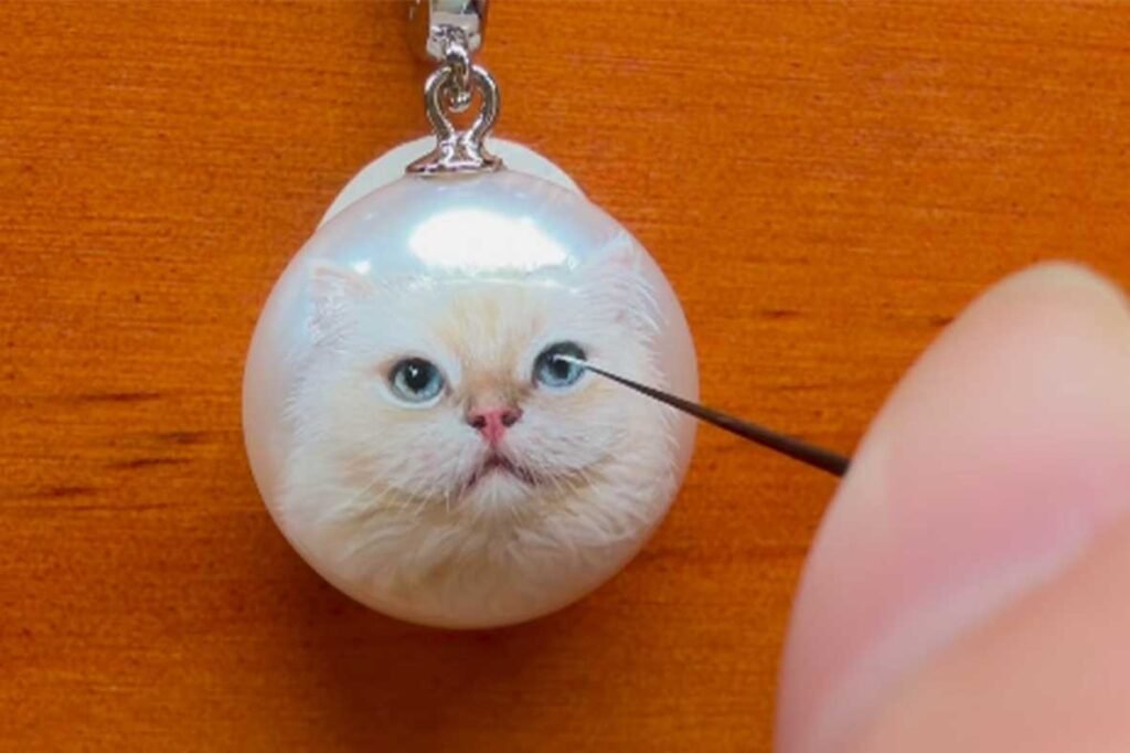 Artisan using a fine needle tool to paint intricate details on a white cat pet portrait necklace, emphasizing craftsmanship.